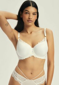 Biały biustonosz z koronkowymi wykończeniami, z formowanymi miseczkami, regulowanymi ramiączkami i zapięciem na haftkę. Gładka faktura z falbaniastymi krawędziami i delikatnymi detalami.