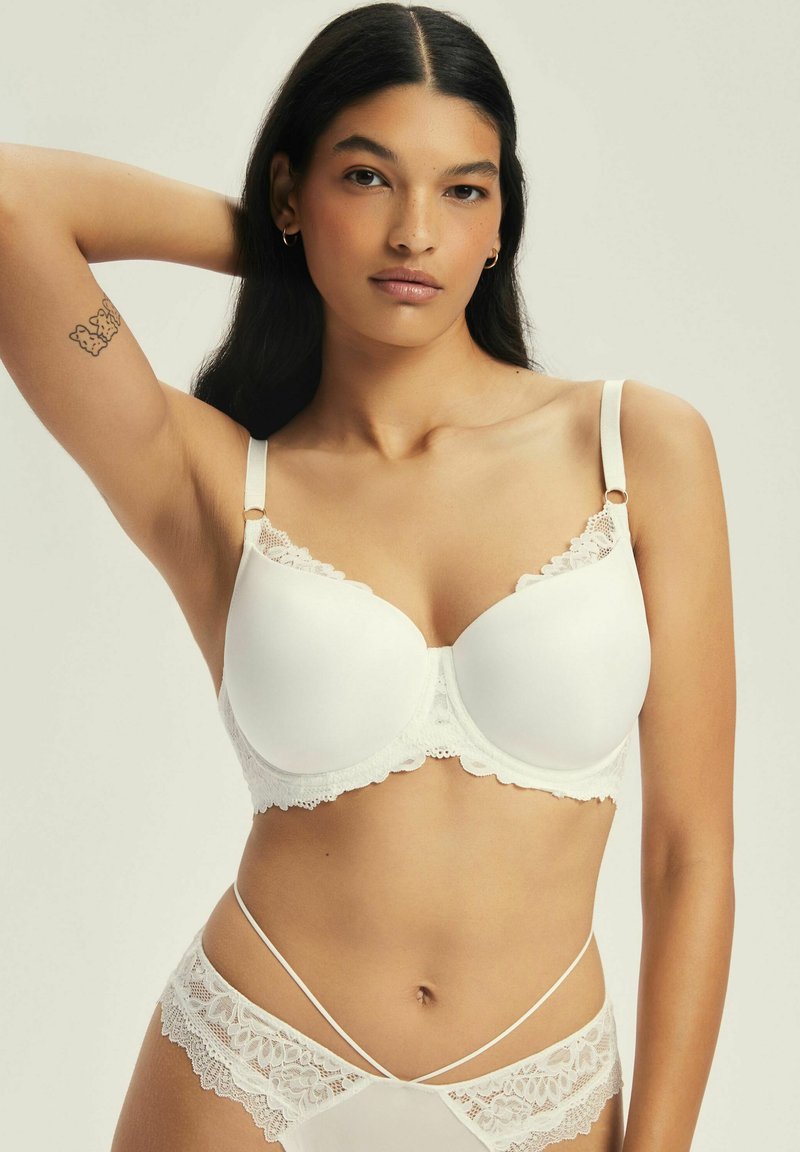 Biały biustonosz z koronkowymi wykończeniami, z formowanymi miseczkami, regulowanymi ramiączkami i zapięciem na haftkę. Gładka faktura z falbaniastymi krawędziami i delikatnymi detalami.