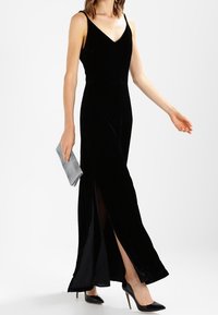Czarna aksamitna sukienka maxi z dekoltem w serek, bocznymi rozcięciami i dopasowaną sylwetką. Modelka trzyma srebrną kopertówkę i nosi czarne szpiczaste szpilki.