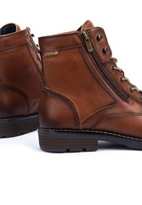 Botas de cuero marrón hasta el tobillo con cremallera y cordones. Cuenta con detalles de costura y una suela texturizada. Etiqueta del fabricante visible.