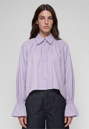 Jeune femme aux longs cheveux foncés portant une chemise lavande ample à boutons avec des manches cloche froncées et un jean sombre taille haute.