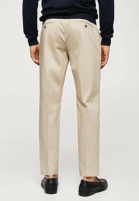 Pantaloni beige su misura con una texture liscia, dotati di due tasche posteriori con chiusura a bottone. Abbinati a scarpe nere.