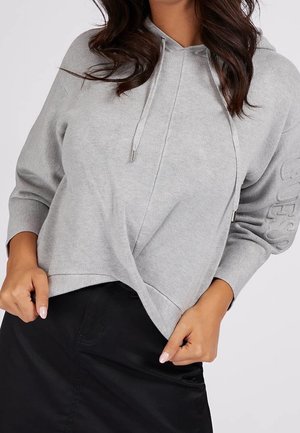 Vrouw draagt een lichtgrijze lange mouwsweater met capuchon, trekkoordjes en een overlappend ontwerp aan de voorkant, gecombineerd met een zwarte rok.