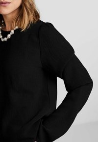 Schwarzer gerippter Pullover mit strukturierter Silhouette, rundem Ausschnitt und puffigen Ärmeln. Ergänzt durch eine chunky Perlenkette.