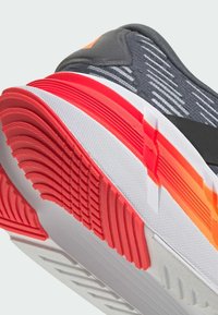 Primo piano di una suola di scarpa atletica nera e grigia con dettagli in rosso e bianco nel battistrada e pattern di presa testurizzati.