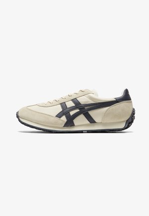 Beige Sportschuh aus Leder und Wildleder, mit dunkelblauen Streifen und einer strukturierten schwarzen Gummisohle. Minimalistisches Design mit Schnürverschluss.