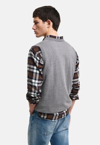 Grå ärmlös tröja över en långärmad skjorta i brunt och blått rutmönster, parat med ljusblå jeans. Enkel design, material i bomullsblandning.