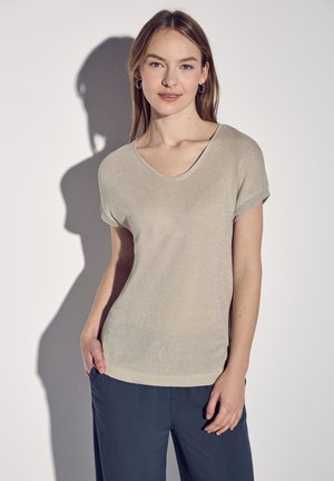 Top beige a maniche corte con scollatura a V, caratterizzato da un leggero luccichio. Il tessuto sembra morbido e leggero, abbinato a pantaloni blu scuro.