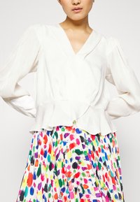 Blusa blanca de manga larga con dobladillo peplum y escote en V. Combinada con una falda plisada de colores y estampados que presenta vibrantes diseños de pinceladas.