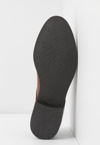 Semelle de chaussure marron avec une surface en caoutchouc noir texturé et une forme courbée, présentant un panneau latéral en cuir marron et une étiquette de taille.