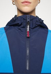 Navy hooded jacket with bright blue panels, featuring a central zipper and branded tag. Smooth texture and lightweight material.
