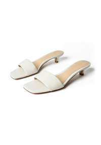 Paire de sandales blanches ouvertes à bout ouvert avec des petits talons aiguilles et des larges brides, placées côte à côte sur un fond blanc.