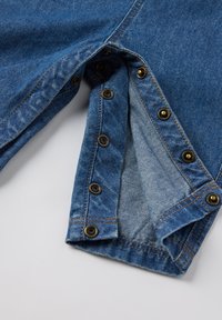 Blauwe denimstof met messing drukknoopjes langs de binnenrand, met een zachte binnenvoering en contrasterende stiksels.