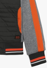 Material de plumas negro con mangas grises; presenta vibrantes detalles en naranja y puños acanalados a rayas. Incluye un parche de marca.