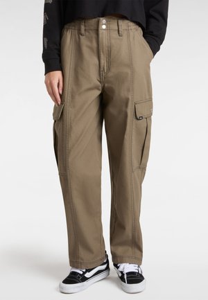 Pantalons cargo beiges à jambes larges, avec poches avant et poches latérales à rabat. Détails cousus et fermeture à bouton en métal. Portés avec des baskets noires.