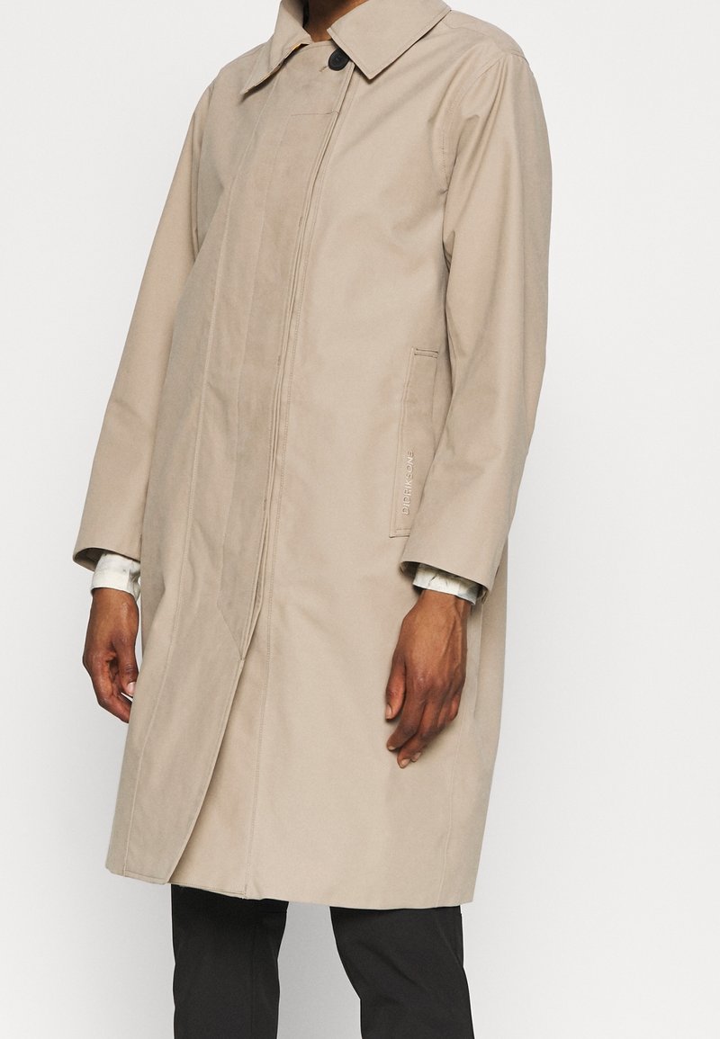 Abrigo largo beige con cuello y bolsillos laterales, usado sobre pantalones negros, mostrando partes de las manos y las mangas contra un fondo blanco.