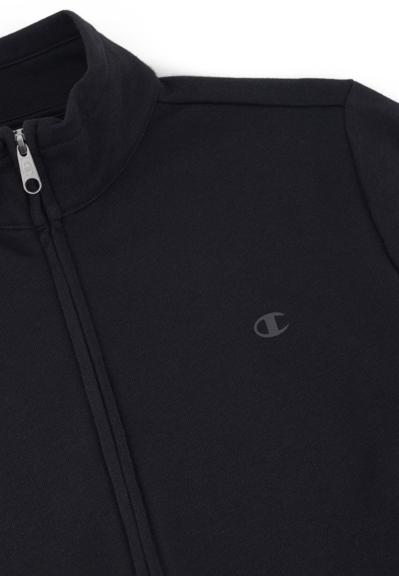 Czarna kurtka zapinana na zamek z wysokim kołnierzem, wykonana z gładkiego materiału. Posiada małe wytłoczone logo na klatce piersiowej i dopasowany krój.