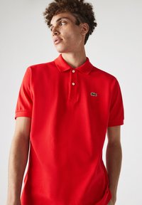 Polo rojo con tejido texturizado, mangas cortas y cuello estándar. Presenta un pequeño logo de cocodrilo verde en el pecho.