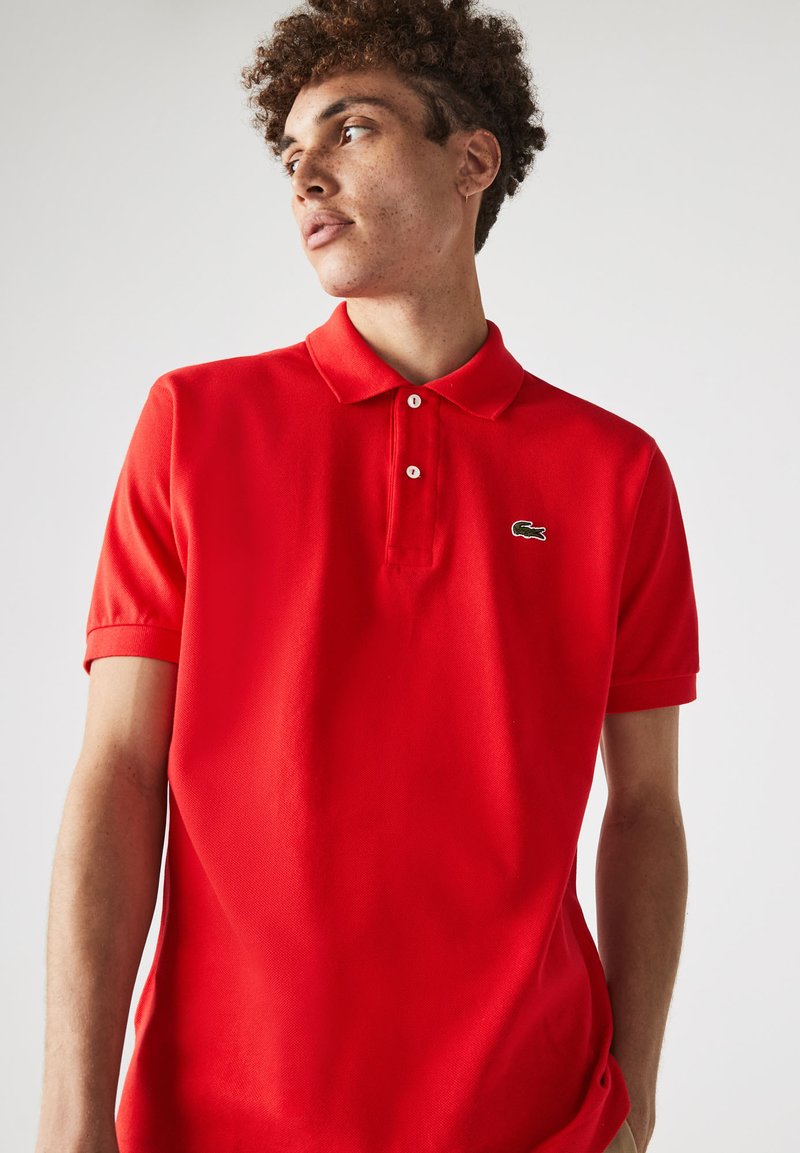 Polo rojo con tejido texturizado, mangas cortas y cuello estándar. Presenta un pequeño logo de cocodrilo verde en el pecho.