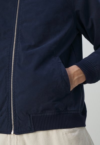 Chaqueta bomber azul marino con cremallera frontal, puños y dobladillo de canalé, y bolsillos laterales. Tejido suave con un ligero brillo y diseño minimalista.