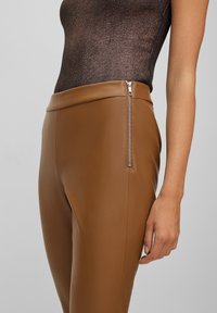 Bruna läderbyxor med en slät textur, som har en sidodragkedja. Midjan är bred och åtsittande, vilket betonar en smickrande silhuett.