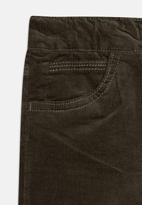 Brūni korduroja pantaloni ar priekšējo kabatu, kas izceļas ar kontrastējošiem šuvumiem, un mīkstu, teksturētu audumu. Jostasvieta ir aprīkota ar elastīgu joslu.