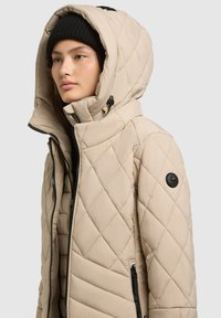 Beige kooti puffer jakk, mis on varustatud kapuutsiga, tõmblukuga, küljetaskutega ja musta logo plaastriga vasakul vikal.