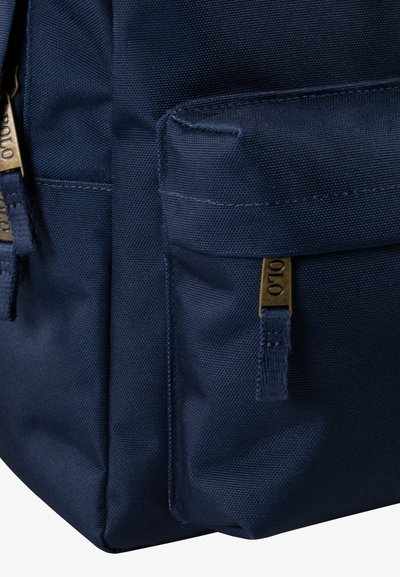 Navy modra teksturirana tkanina nahrbtnik z dvema sprednjima žepoma na zadrgo in bronastimi kovinskimi dodatki. Robustna zasnova primerna za vsakodnevno uporabo.