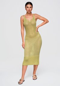 Femme aux cheveux foncés portant une robe midi tricotée sans manches vert clair et des sandales, debout avec une main sur la hanche devant un fond uni.