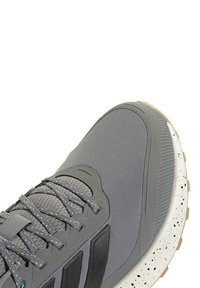 Zapatilla deportiva gris con parte superior de tejido texturizado, cordones elásticos y una suela blanca moteada. Presenta rayas negras en el costado.