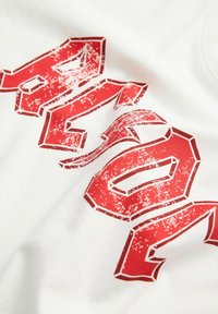Hvid bomulds t-shirt med fed, slidt rød tekst i et omridsdesign. Nært blik på grafikkens placering og tekstur.