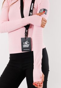Maglietta a maniche lunghe rosa chiaro con texture a coste e design aderente, abbinata a leggings neri. Presenta un laccetto e un'etichetta del logo sul petto.