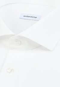 Chemise blanche en tissu texturé, avec un col classique et un bouton blanc unique. Étiquette indiquant "seidensticker".