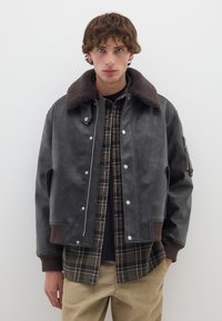 Giacca bomber in pelle nera con colletto in pile marrone, bottoni a pressione argentati e tasca laterale con zip, indossata sopra una camicia a quadri.