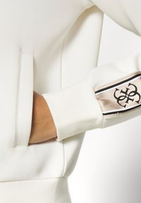 Hvit sweatshirt med ribbestrikkede mansjetter, prydet med en beige satenglapp med sort brodert logo og en skjult sidelomme.