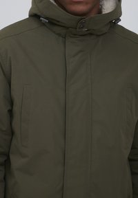 Olivgrön huva parkas tillverkad av texturerat tyg med framdragkedja och knappar, med sidofickor och mjukt fleecefoder.