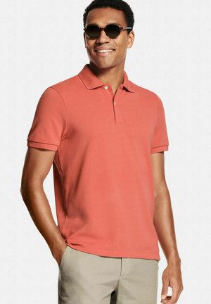 Lachende man met donkere zonnebril, een koraalrood poloshirt met korte mouwen en beige broek, met één hand in de zak tegen een effen achtergrond.