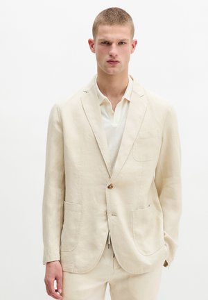 Junger Mann trägt einen cremefarbenen Leinenanzug mit passender Hose und weißem Hemd, steht vor einem schlichten weißen Hintergrund.