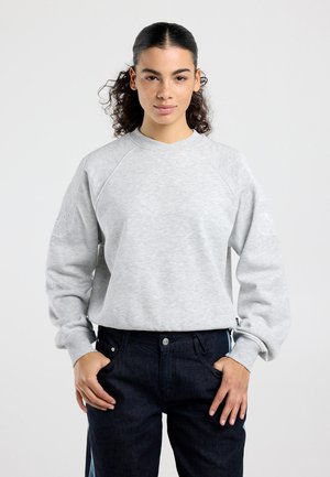 Jeune personne aux cheveux bouclés attachés en queue de cheval portant un sweat-shirt gris clair et un jean foncé, debout avec les mains partiellement dans les poches.