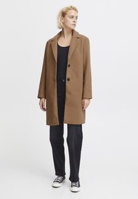 Cappotto di lana marrone con colletto a punta, due bottoni e taglio dritto, abbinato a jeans neri e sneaker stringate.
