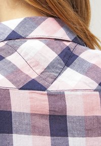 Set bagfra af en person iført en lyserød, marineblå og hvid ternet skjorte med synlige syninger og skuldersøm, hår delvist dækker nakken.