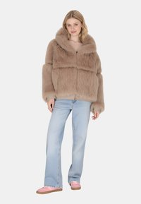 Giacca in pelliccia sintetica di un morbido beige con un ampio colletto, vestibilità oversize e maniche lunghe; indossata sopra una maglietta nera e abbinata a jeans chiari e scarpe rosa.