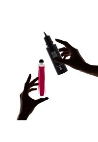 Due mani che tengono una bottiglia di profumo trasparente rosa e una bottiglia spray nera. Lo sfondo è bianco, enfatizzando le forme e i colori dei prodotti.