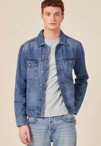 Vaate denim takki vaaleansinisena, kahdella rintataskulla, nappikiinnityksellä ja klassisella kauluksella. Päällä vaalea harmaa T-paita ja farkut.