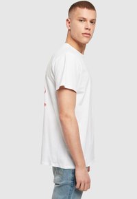 Weißes Baumwoll-T-Shirt mit kurzen Ärmeln, lockerer Passform, mit rotem Grafiktext auf der Rückseite. Einfacher Ausschnitt und glatte Textur.