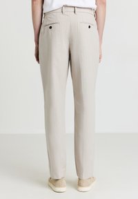Pantalon beige à rayures fines avec une coupe ajustée, comportant des poches arrière avec des boutons noirs et une texture de tissu lisse.