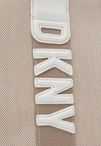 Sac en tissu beige avec une surface texturée et un grand lettrage DKNY blanc. Le design comprend des accents cousus et une bandoulière en cuir lisse.