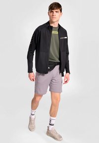 Svart idrottsjacka med hög krage, i kombination med en grön kortärmad tröja och grå shorts. Kompletta med beige sneakers och randiga strumpor.