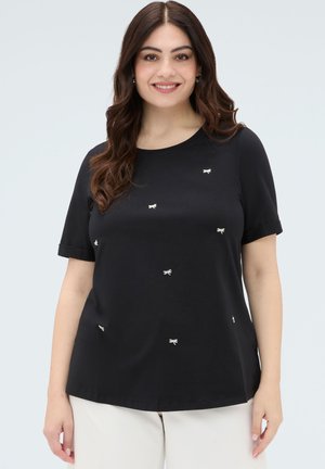 Mujer sonriente con cabello largo y castaño, que lleva una camiseta negra de manga corta con pequeños dibujos de lazos blancos y pantalones de color claro.