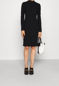 Robe noire à manches longues avec un col montant, une silhouette ajustée et une texture lisse. Associée à un sac fourre-tout blanc et des mocassins noirs ornés d'une chaîne dorée.